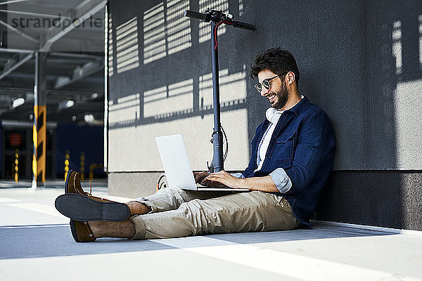 Lächelnder junger Mann mit Elektroroller sitzt mit Laptop auf dem Boden