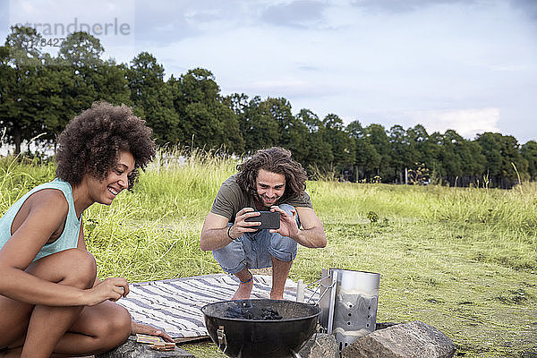 Lächelnder junger Mann mit Freundin beim Fotografieren eines Barbecue-Grills in der Natur