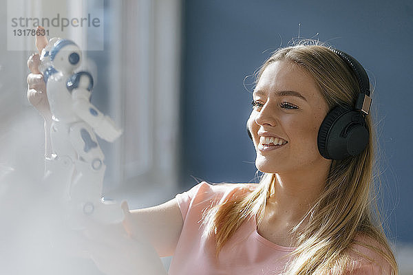 Porträt einer lächelnden jungen Frau mit Kopfhörern  die einen Spielzeugroboter ansieht