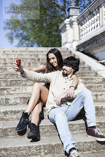 Glückliches junges Paar sitzt auf der Treppe und macht sich mit dem Smartphone selbstständig