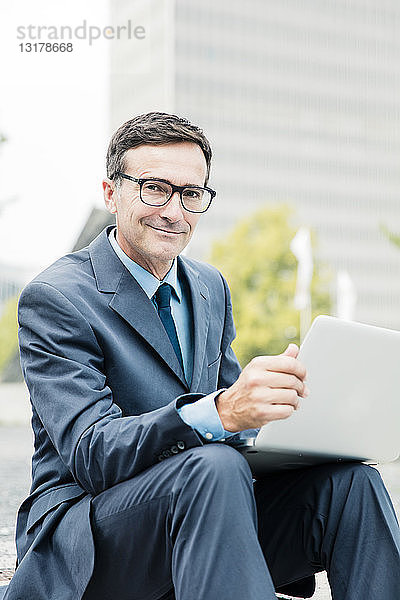 Porträt eines lächelnden Geschäftsmannes  der in der Stadt am Laptop sitzt