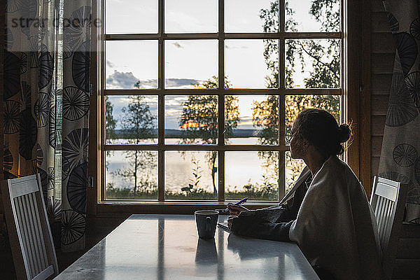 Finnland  Lappland  junge Frau sitzt am Fenster und schaut auf einen See