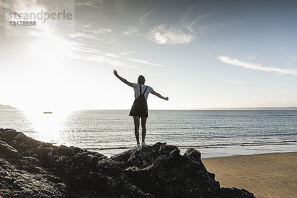Frankreich  Bretagne  Rückansicht einer jungen Frau  die bei Sonnenuntergang auf einem Felsen am Strand steht