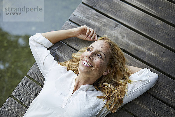 Lächelnde blonde Frau liegt auf einem Holzsteg an einem See