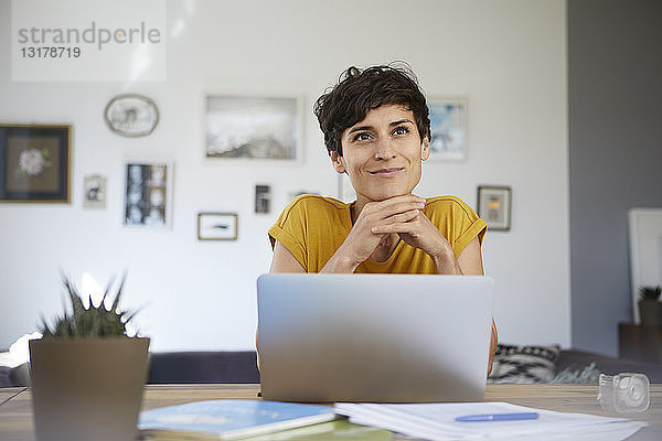 Porträt einer lächelnden Frau  die zu Hause am Tisch sitzt und einen Laptop benutzt