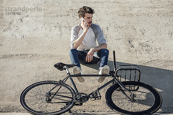 Junger Mann mit Pendler-Fixie-Fahrrad sitzt auf Betonwand und telefoniert