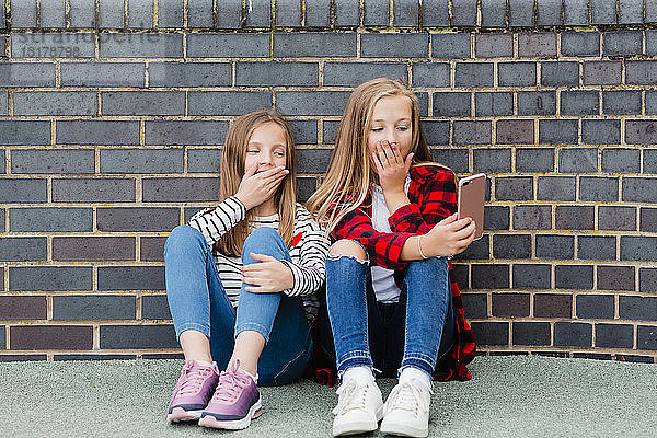 Porträt von zwei Mädchen  die vor einer Ziegelmauer sitzen und sich mit einem Smartphone selbstständig machen