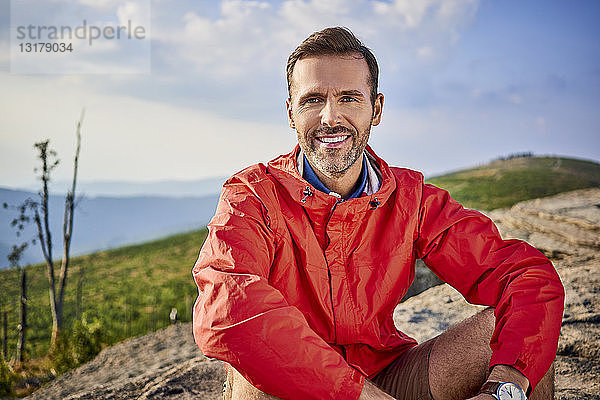 Porträt eines lächelnden Mannes  der während einer Wanderung auf einem Felsen sitzt