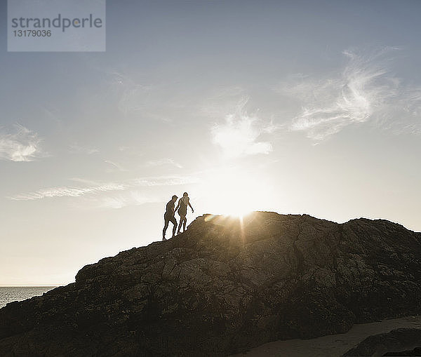 Frankreich  Bretagne  junges Paar klettert bei Sonnenuntergang auf einen Felsen am Strand