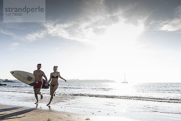 Frankreich  Bretagne  glückliches junges Paar mit einem Surfbrett  das Hand in Hand im Meer läuft