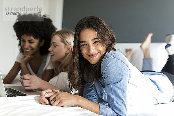 Porträt einer lächelnden jungen Frau  die mit Freundinnen am Bett liegt und einen Laptop benutzt
