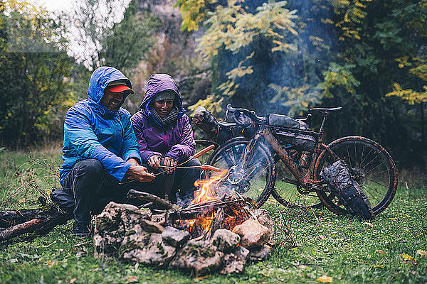 Lachendes Paar mit bmx-Fahrrädern sitzt am Lagerfeuer und grillt Würstchen