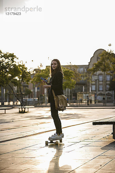 Junge Frau mit Handy auf Longboard in der Stadt
