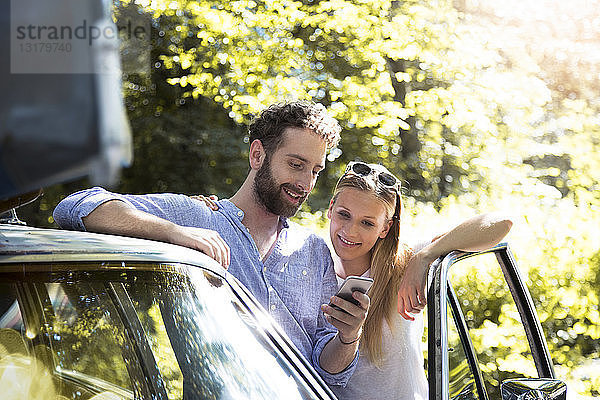 Lächelndes junges Paar mit Handy am Auto im Wald