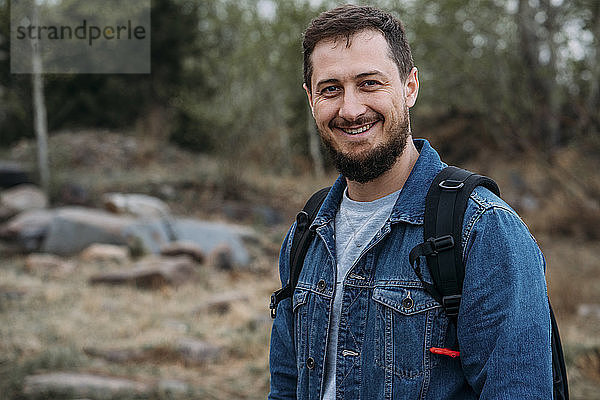 Porträt eines lächelnden Mannes mit Rucksack und Jeansjacke in Natur
