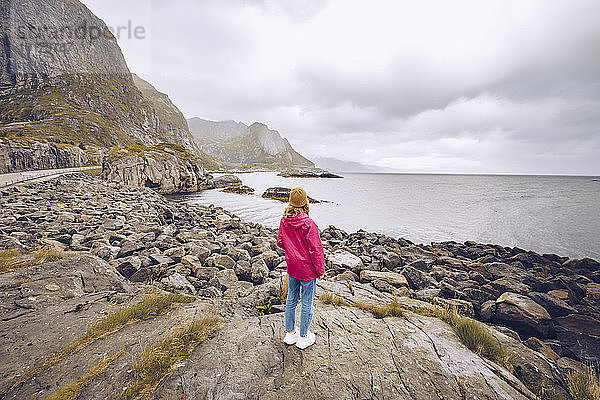 Norwegen  Lofoten  Rückenansicht einer jungen Frau in Regenjacke  die auf einem Felsen steht und in die Ferne schaut