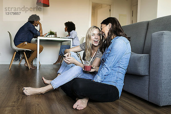 Zwei fröhliche junge Frauen sitzen auf dem Boden mit Handy und Getränken und Freunden im Hintergrund
