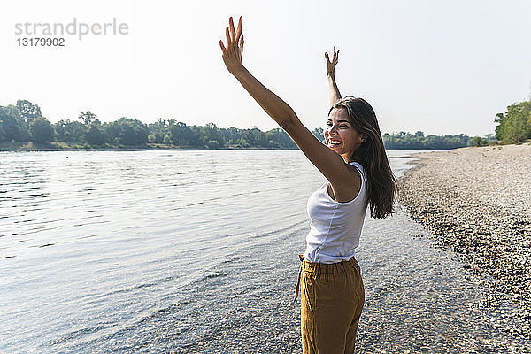 Glückliche junge Frau steht am Flussufer und hebt ihre Arme