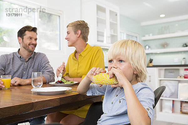 Junge isst Maiskolben in der Küche mit den Eltern im Hintergrund