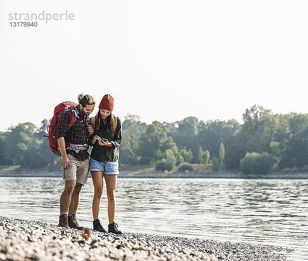 Junges Paar mit Rucksäcken am Flussufer beim Handy-Check