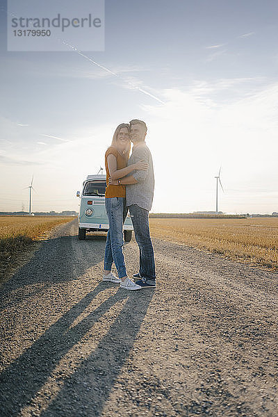 Zärtliches junges Paar steht auf Feldweg am Wohnmobil in ländlicher Landschaft
