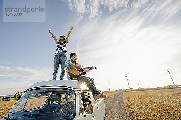 Glückliches Paar mit Gitarre auf dem Dach eines Wohnmobils in ländlicher Landschaft