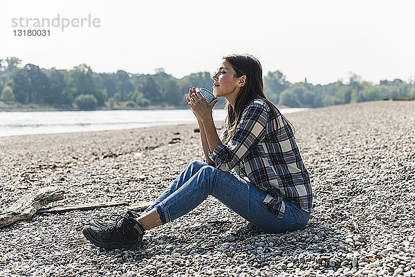 Entspannte junge Frau  die am Flussufer sitzend einen Becher hält