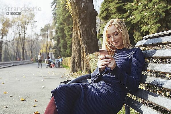 Porträt einer blonden Frau  die auf einer Bank im herbstlichen Stadtpark sitzt und ein Smartphone benutzt
