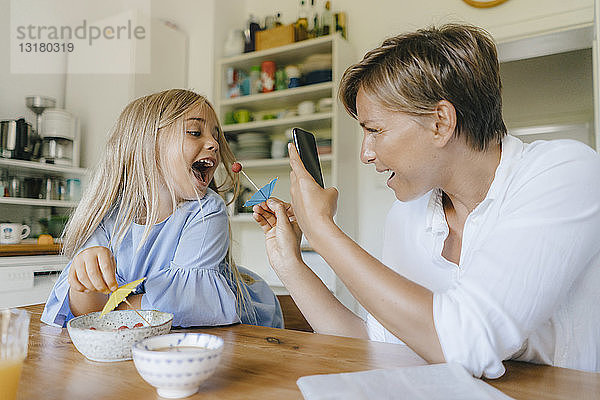 Glückliche Mutter und Tochter amüsieren sich zu Hause am Tisch und fotografieren mit dem Smartphone