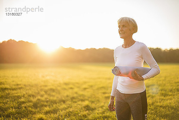 Ältere Frau hält Yogamatte bei Sonnenuntergang auf einer ländlichen Wiese