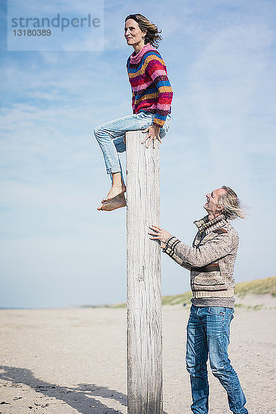 Reifer Mann hilft Frau beim Klettern auf einen Holzpfahl am Strand