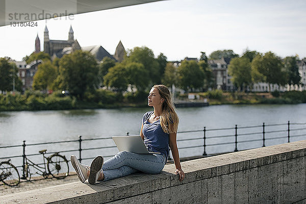Niederlande  Maastricht  junge Frau sitzt mit Laptop auf einer Mauer am Flussufer