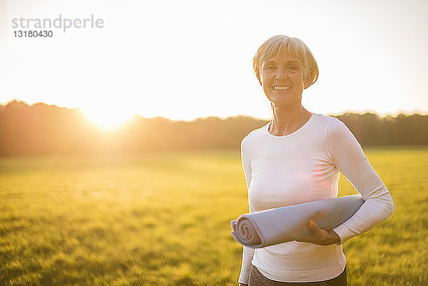 Porträt einer lächelnden älteren Frau  die bei Sonnenuntergang auf einer ländlichen Wiese eine Yogamatte hält