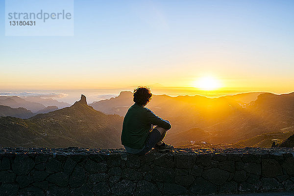 Spanien  Kanarische Inseln  Gran Canaria  Rückenansicht eines Mannes  der auf einer Mauer sitzt und den Sonnenuntergang über einer Berglandschaft betrachtet