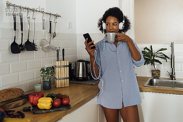 Frau mit Kopfhörern  die ein Smartphone benutzt und Kaffee zum Frühstück in ihrer Küche trinkt