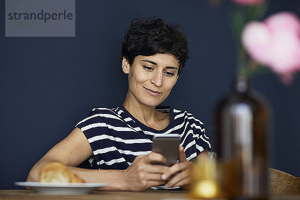 Lächelnde Frau zu Hause  die am Holztisch sitzt und ihr Handy kontrolliert