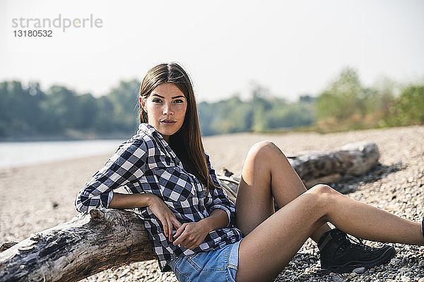 Junge Frau sitzt am Kieselsteinufer am Baumstamm