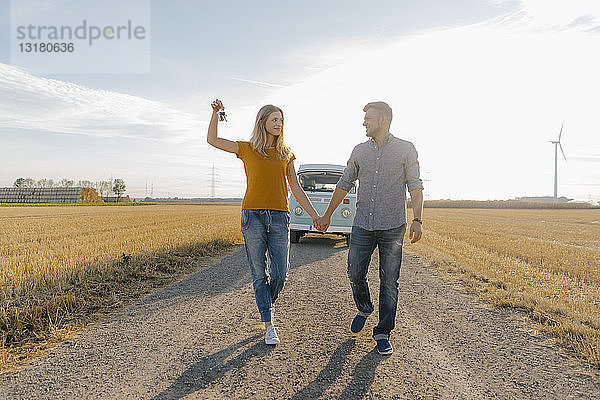 Junges Paar mit Autoschlüssel zu Fuß auf Feldweg mit Wohnmobil in ländlicher Landschaft