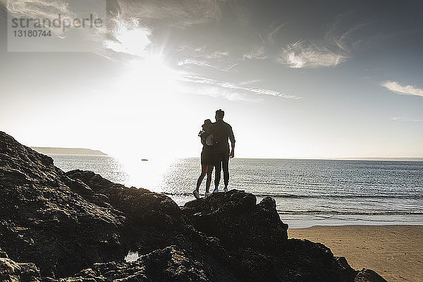 Frankreich  Bretagne  Rückansicht eines jungen Paares  das bei Sonnenuntergang auf einem Felsen am Strand steht