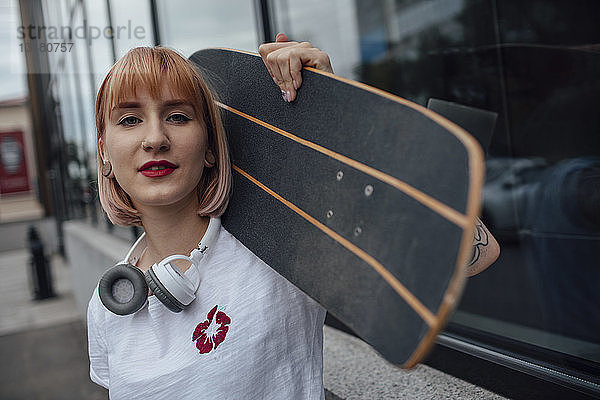 Porträt einer selbstbewussten jungen Frau  die ein Carver-Skateboard vor einem Gebäude hält