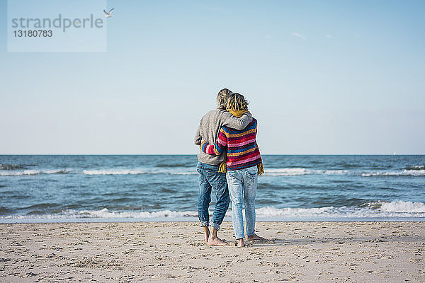 Ein Paar  das mit den Armen am Strand steht und auf das Meer schaut