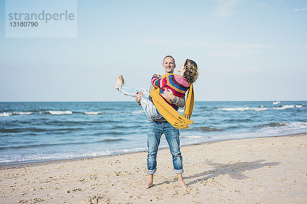 Glücklicher Mann trägt seine Frau auf dem Arm am Strand