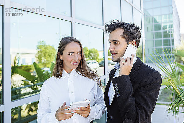 Lächelnde Geschäftsfrau und Geschäftsmann mit Tablet und Handy vor dem Bürogebäude