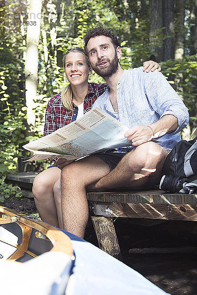 Junges Paar mit Karte und Kanu auf einem Steg an einem Waldbach sitzend