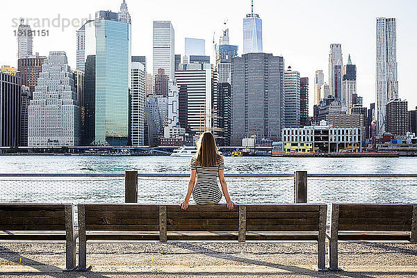 USA  New York  Brooklyn  Rückenansicht einer Frau  die auf einer Bank vor dem East River und der Skyline von Manhattan sitzt