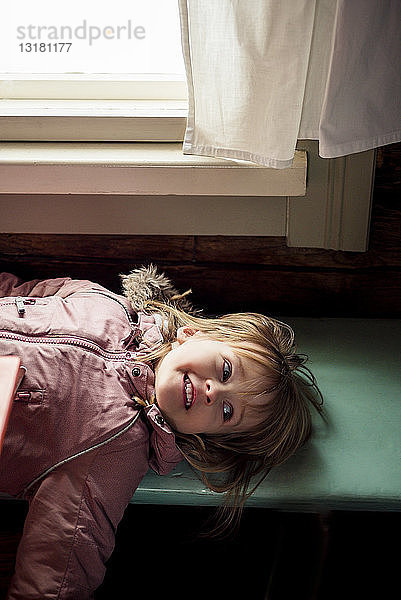 Porträt eines lächelnden kleinen Mädchens  das auf einer Holzbank liegt und im Haus einen Anorak trägt