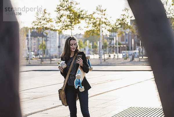 Junge Frau mit Longboard und Snack in der Stadt beim Handy-Check