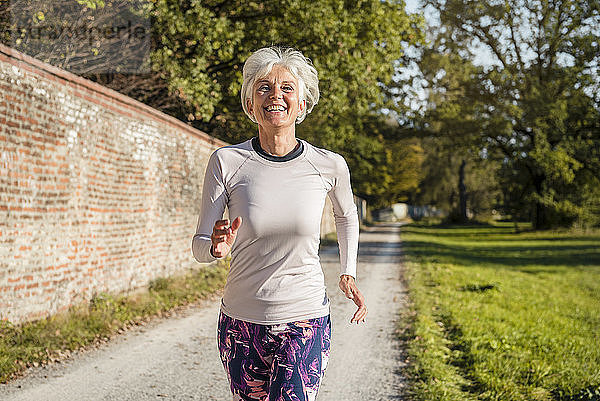 Glückliche ältere Frau läuft in einem Park an einer Ziegelmauer entlang