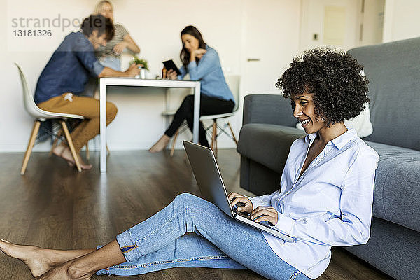 Lächelnde Frau sitzt auf dem Boden und benutzt einen Laptop mit Freunden im Hintergrund