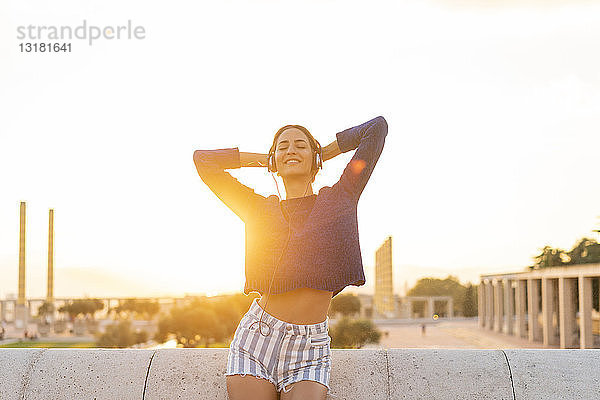Spanien  Barcelona  Montjuic  glückliche junge Frau  die bei Sonnenuntergang mit Kopfhörern Musik hört